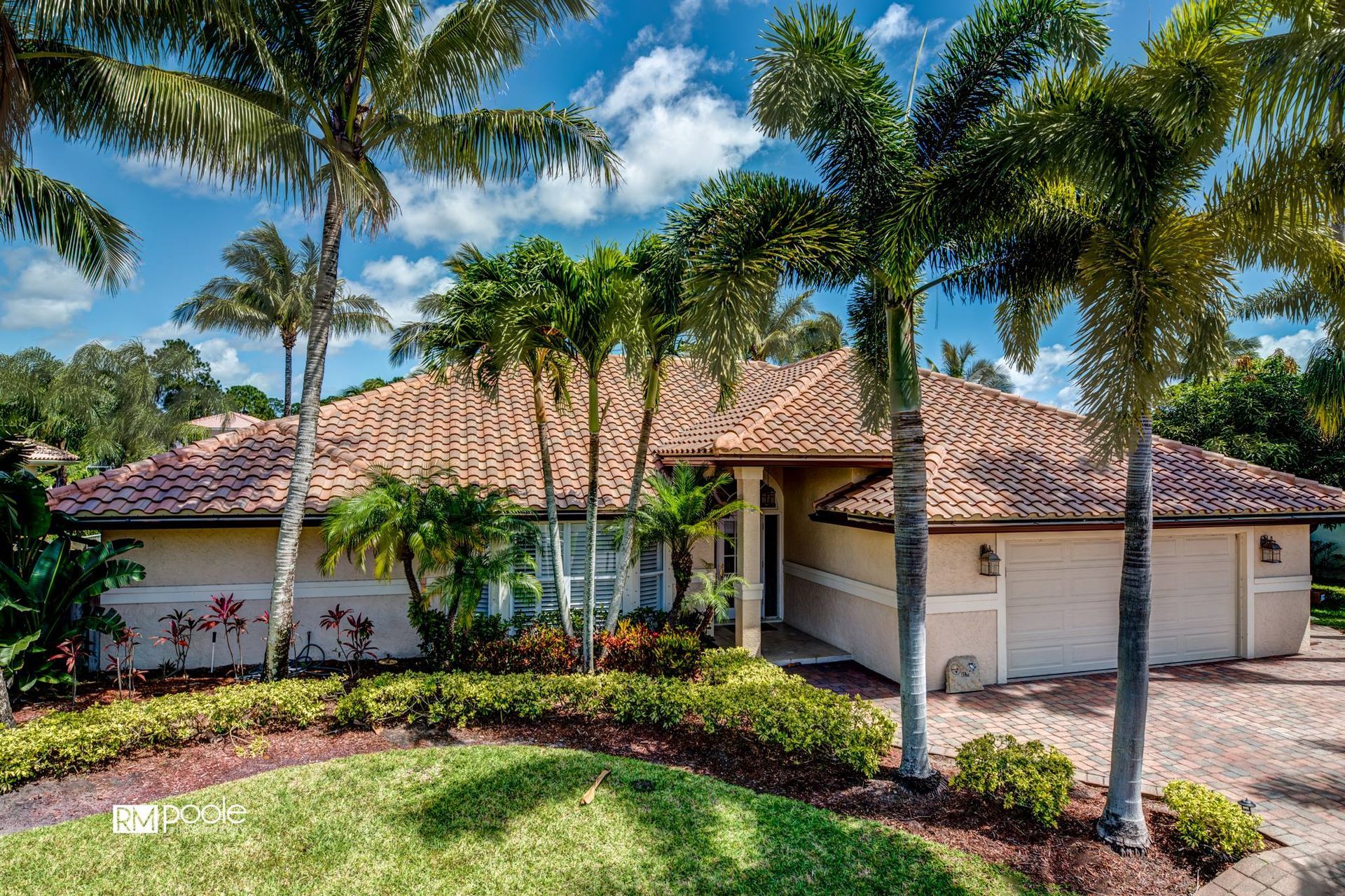 A tan house with a brown tiled roof and palm trees in the front yard under a blue sky with scattered clouds.