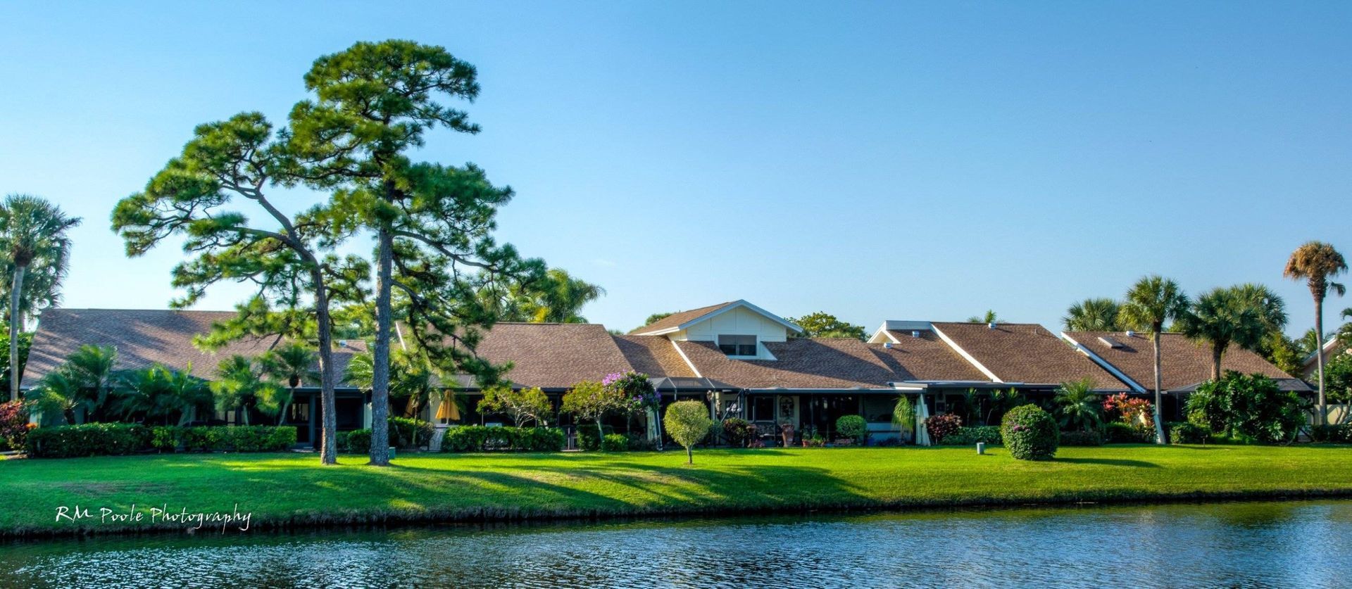 Row of single-story houses with brown roofs and lush green lawns bordering a calm canal under a clear blue sky.
