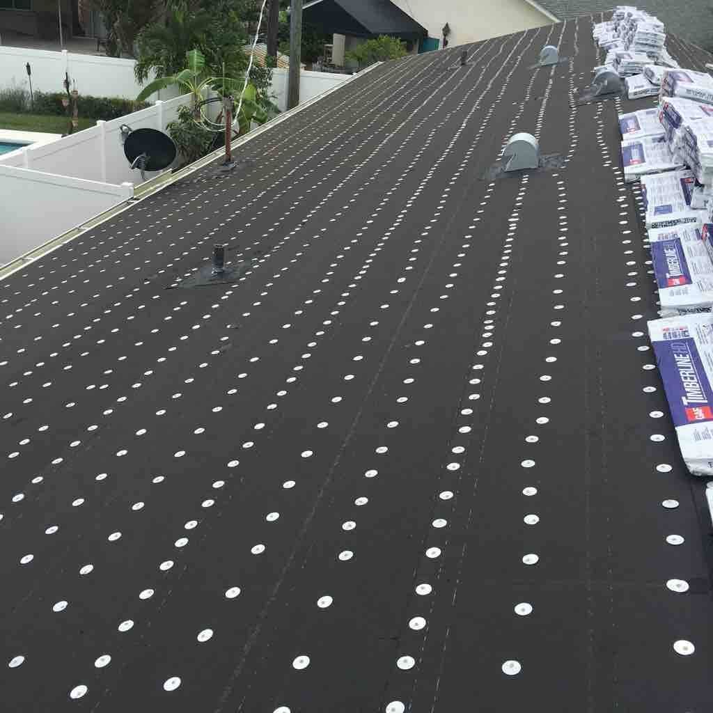 Roof underlayment installed on a sloped residential roof with many white fasteners, next to stacks of roofing materials.