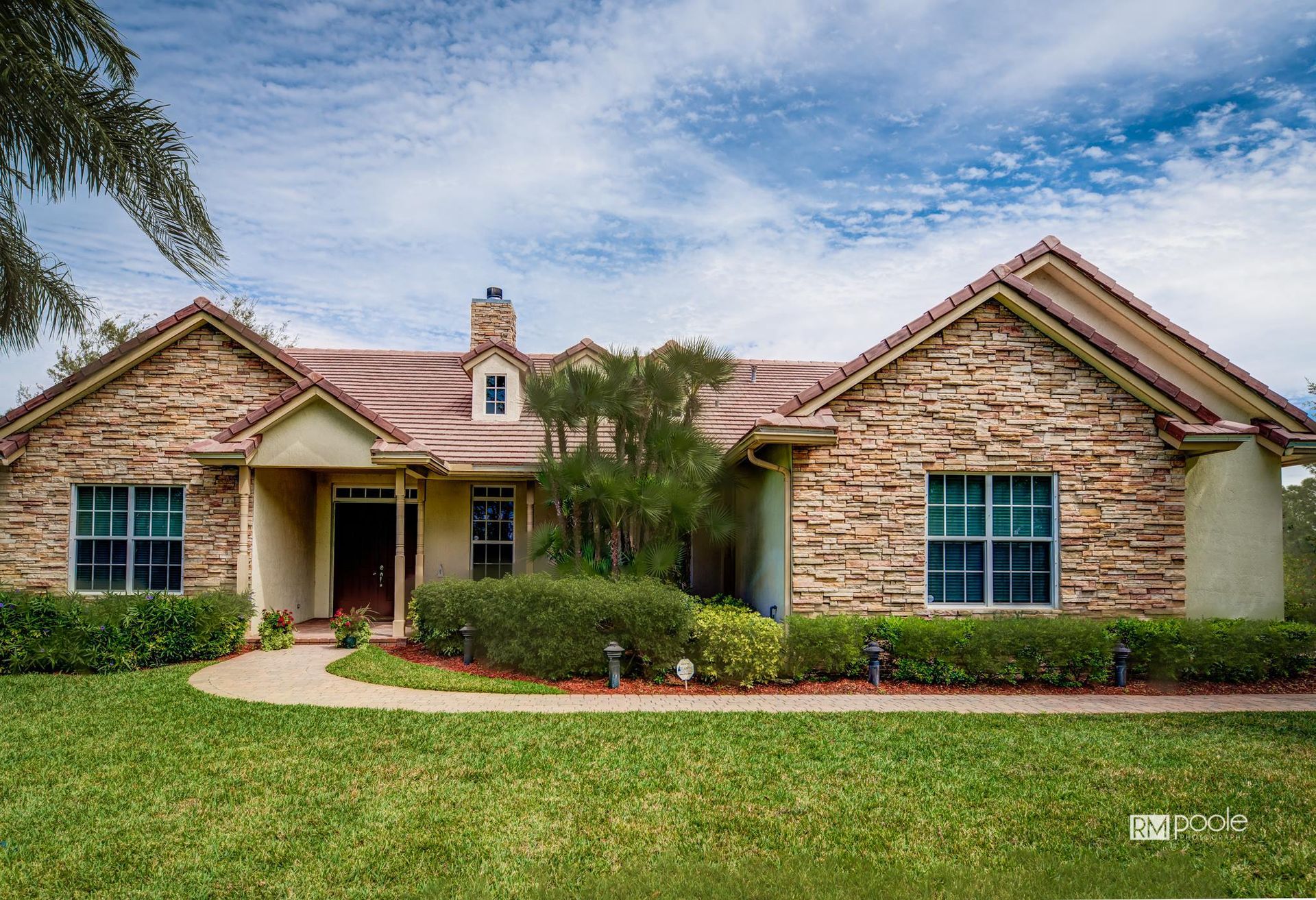 A single-story stone-faced house with a red tile roof, surrounded by green landscaping under a bright blue sky.