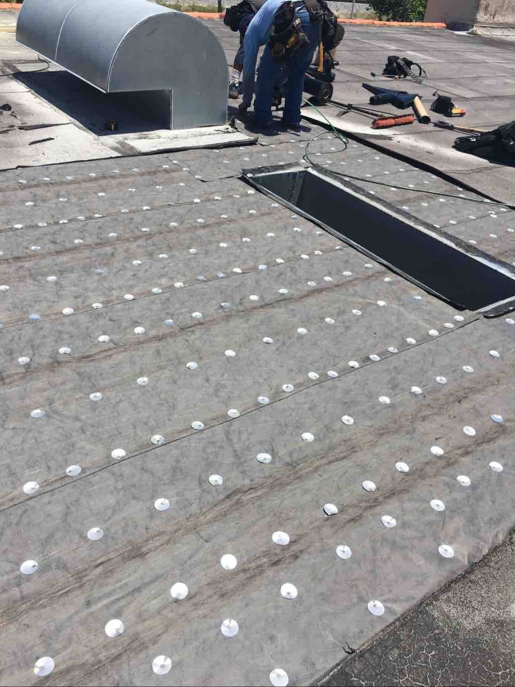 A worker installs roofing material featuring a grid of circular fasteners on a flat commercial roof with an open hatch.