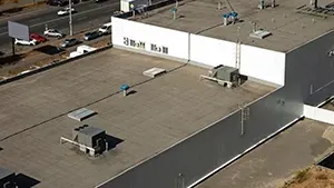 Aerial view of a flat commercial building roof with HVAC units, vents, and a neighboring road with parked cars.