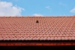 A section of a terracotta-colored tiled roof against a clear blue sky with a single circular roof vent.