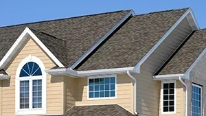 The top exterior of a two-story house with beige siding, white trim, and a shingled roof under a clear blue sky.