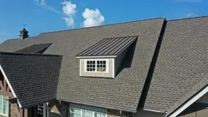 A low-angle view of a grey shingled house roof featuring a metal-roofed dormer window against a bright blue sky.
