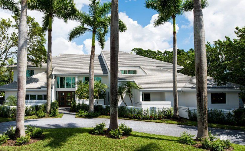 A multi-story, white house with a gray roof, surrounded by tall palm trees and lush greenery under a partly cloudy sky.