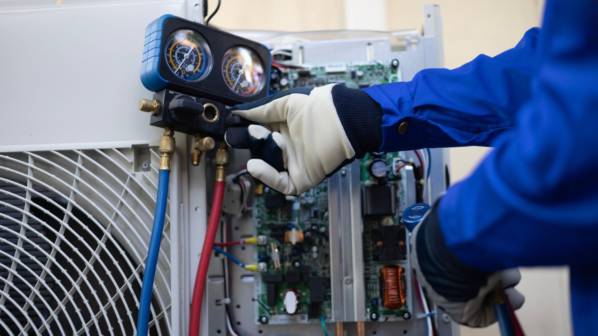 A person in a blue jumpsuit and gloves repairs HVAC equipment using gauges and hoses.