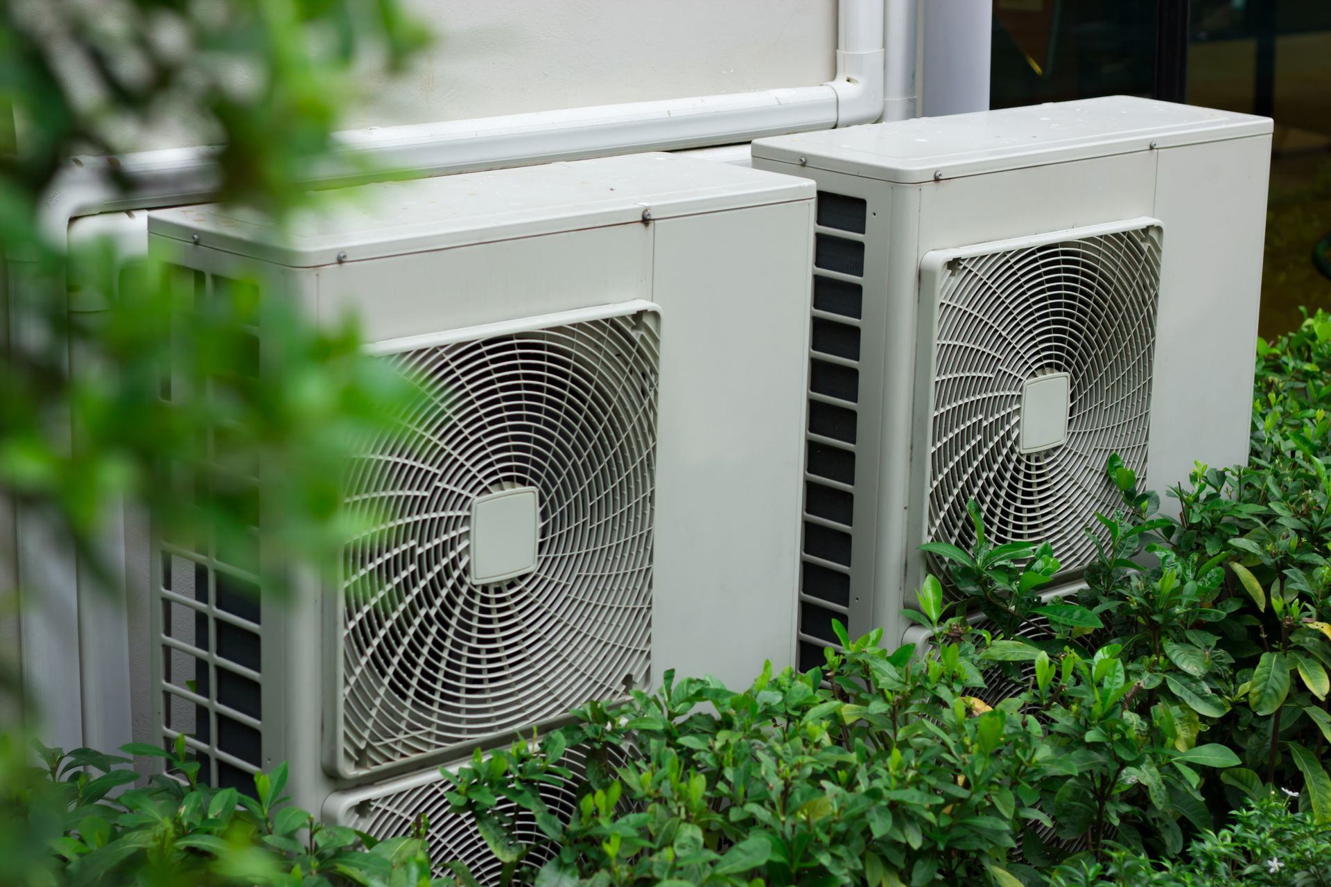 Two white air conditioning units partially obscured by green bushes.