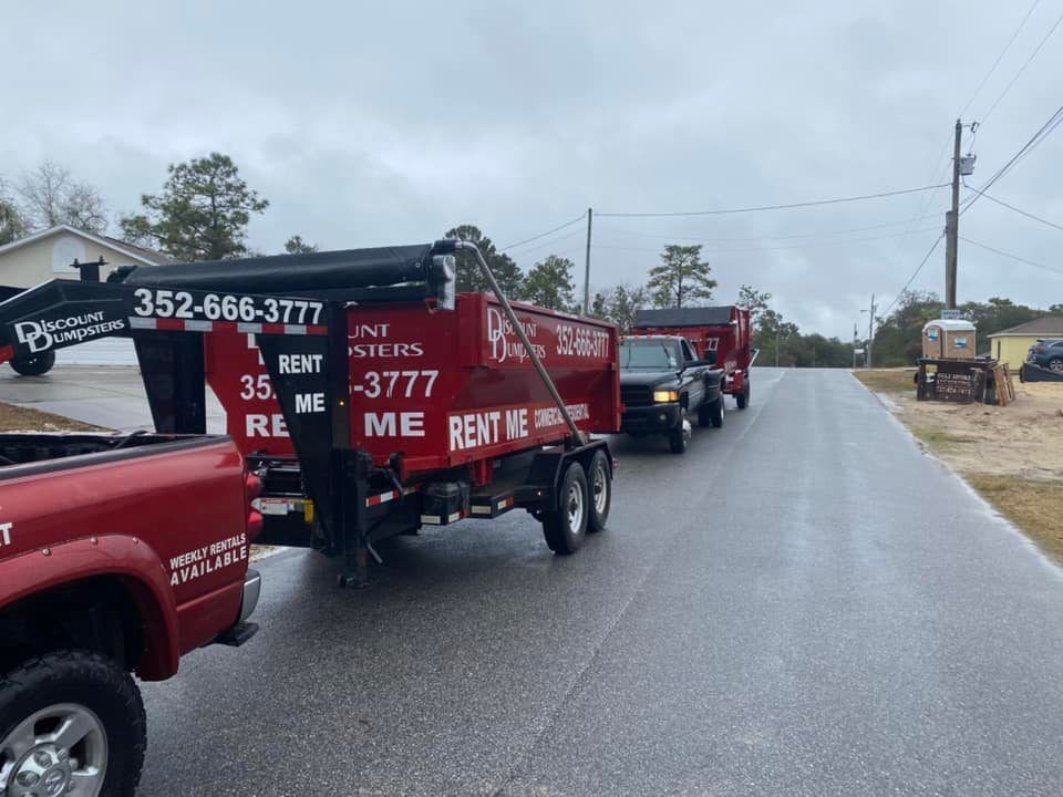 A red truck is driving down a road next to a dumpster.