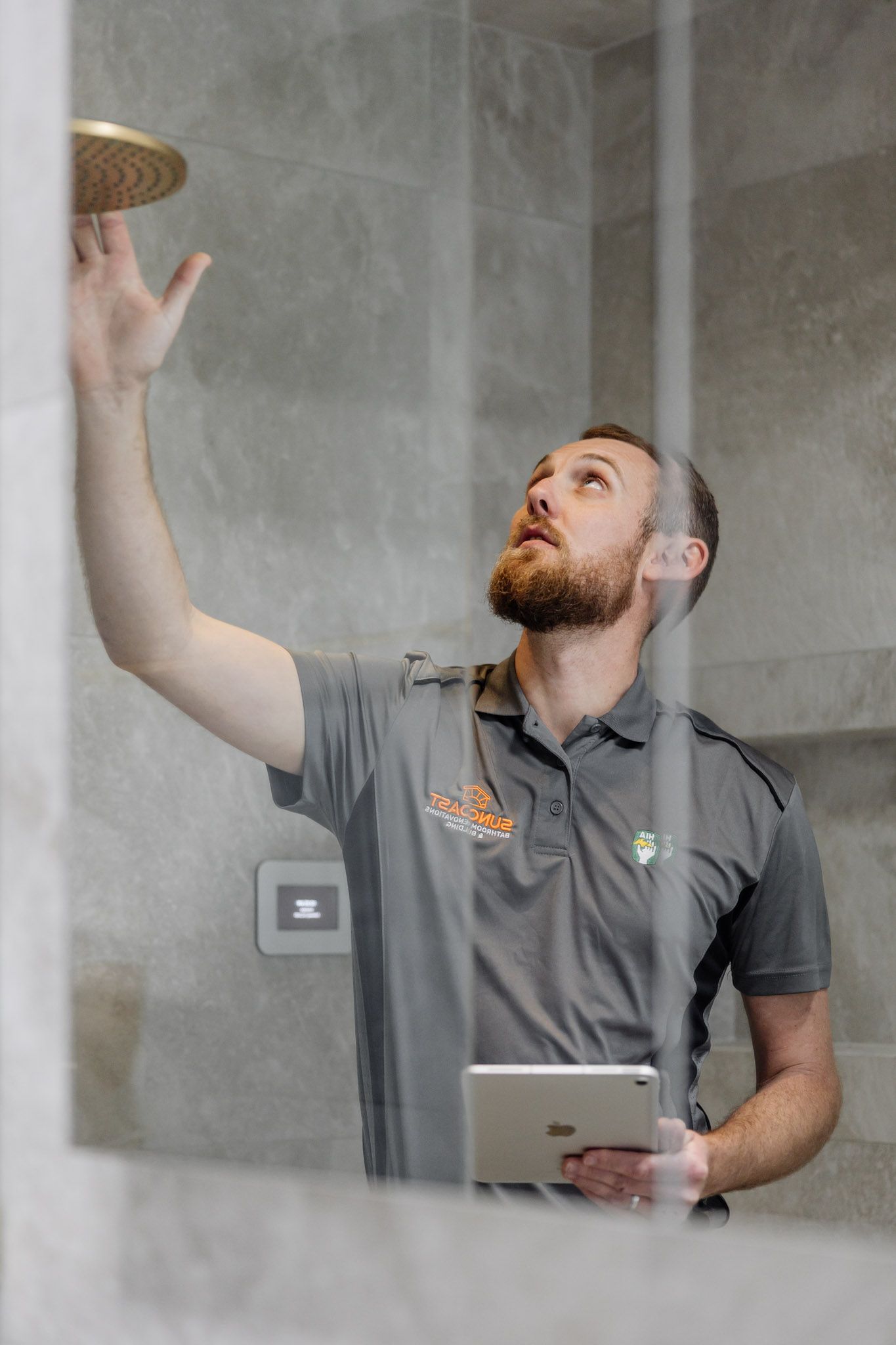 Man in gray shirt inspecting showerhead, holding tablet. Inside a shower with glass wall.