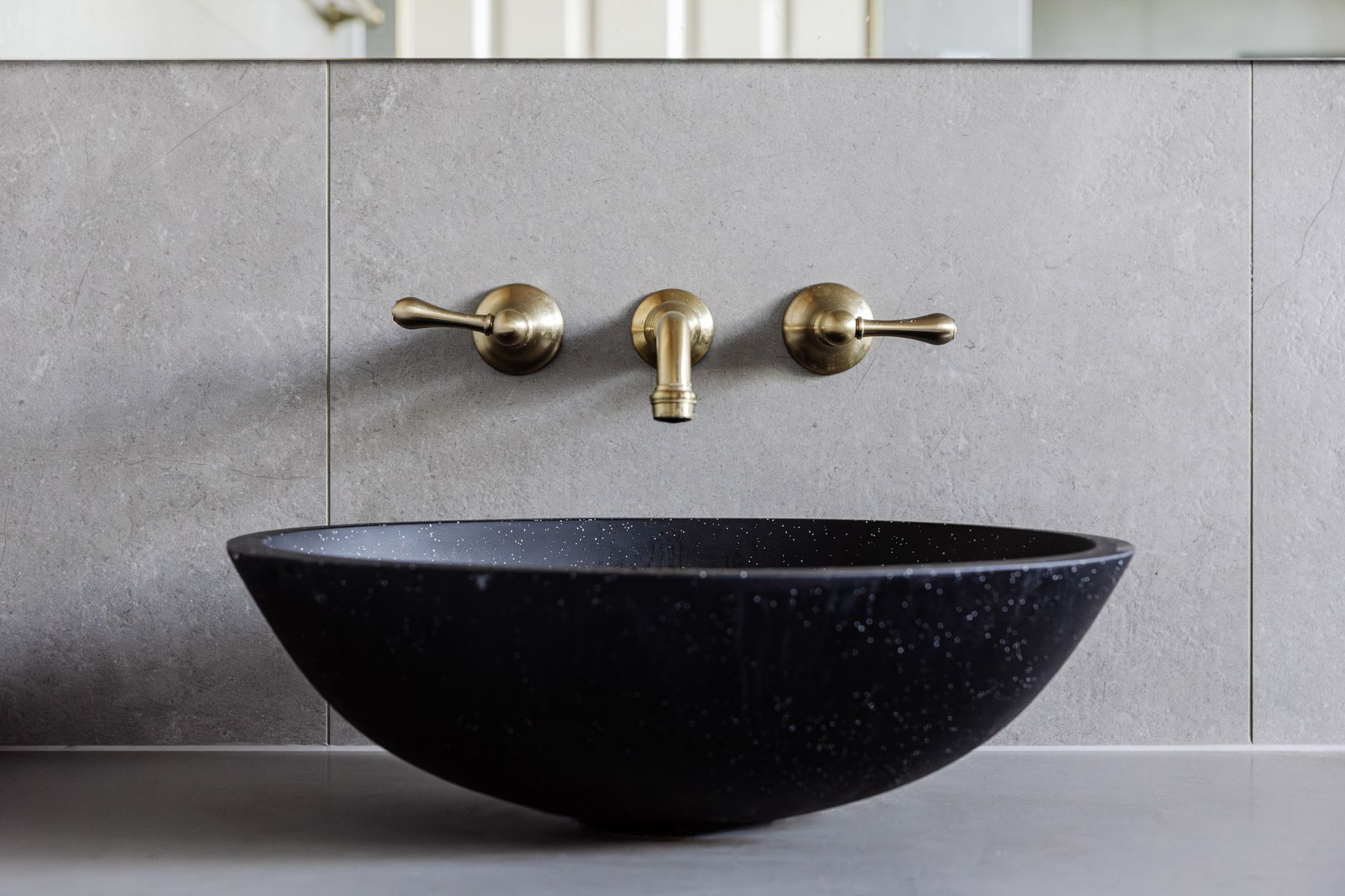 Black speckled vessel sink with brass faucet against a gray tile wall.