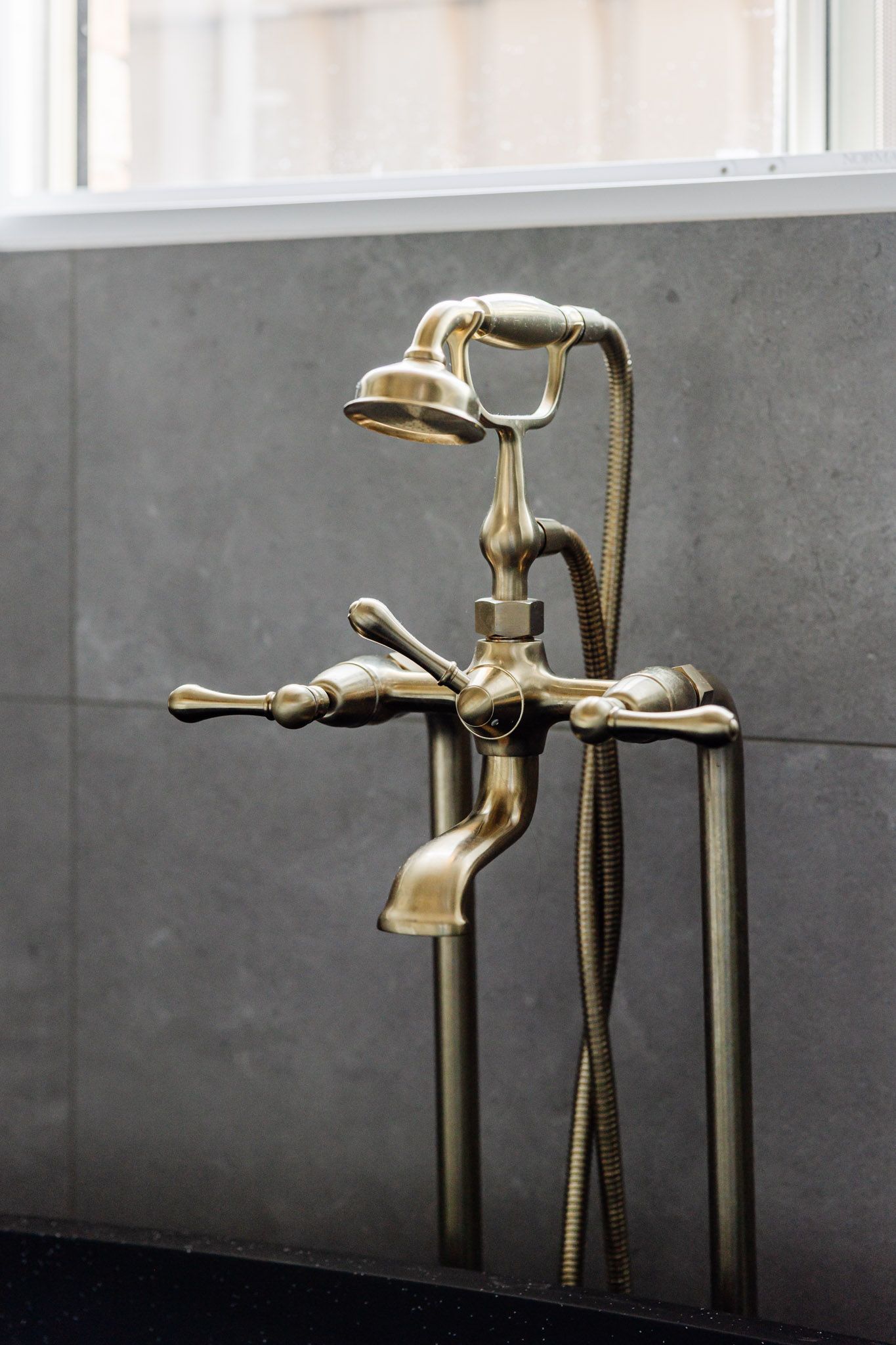 Antique brass bathtub faucet with a shower head, against a gray wall.