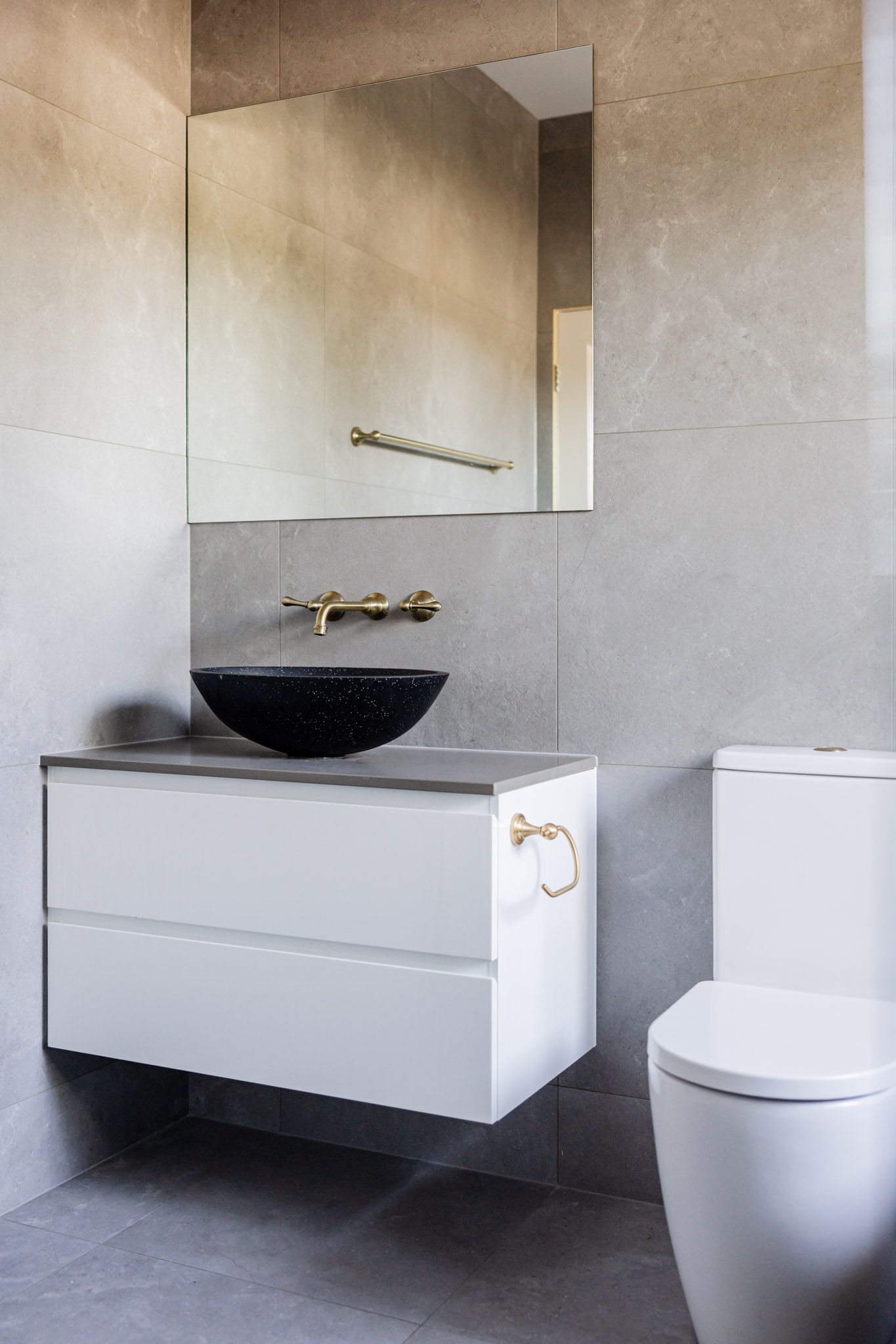 Modern bathroom with gray tiles, floating white vanity, black sink, gold fixtures, and a toilet.