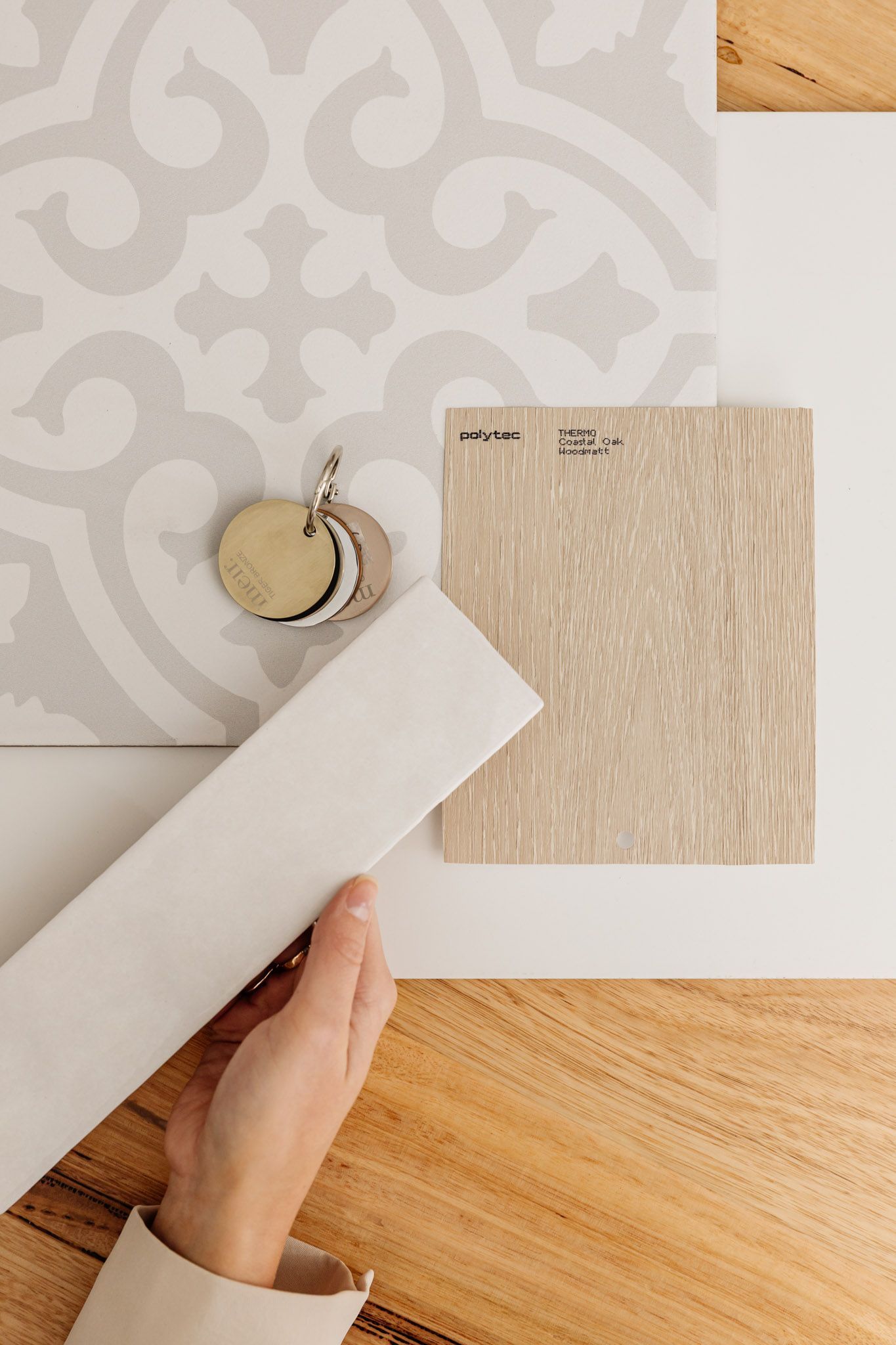 Hand holding a white fabric strip next to a patterned wallpaper, gold textured sample, and wood surface.