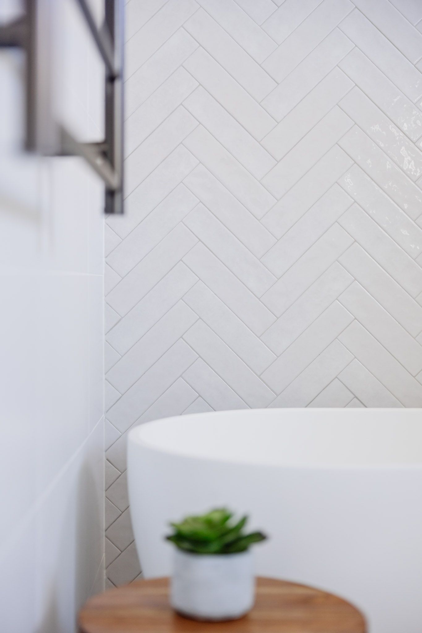 White herringbone tile bathroom wall, rounded bathtub, and potted succulent on wooden stool.