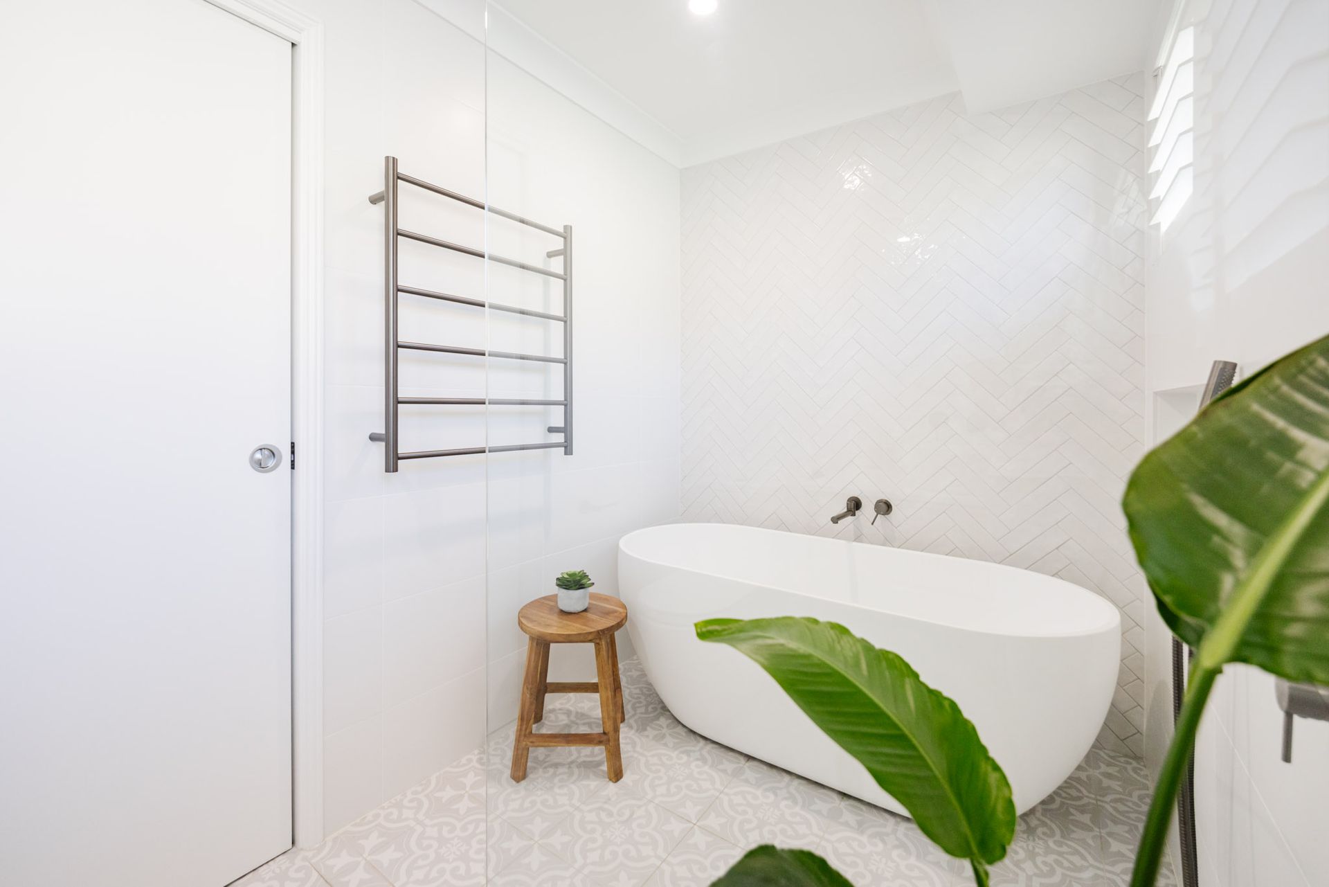 White bathroom with a freestanding tub, towel warmer, and wooden stool with a plant.