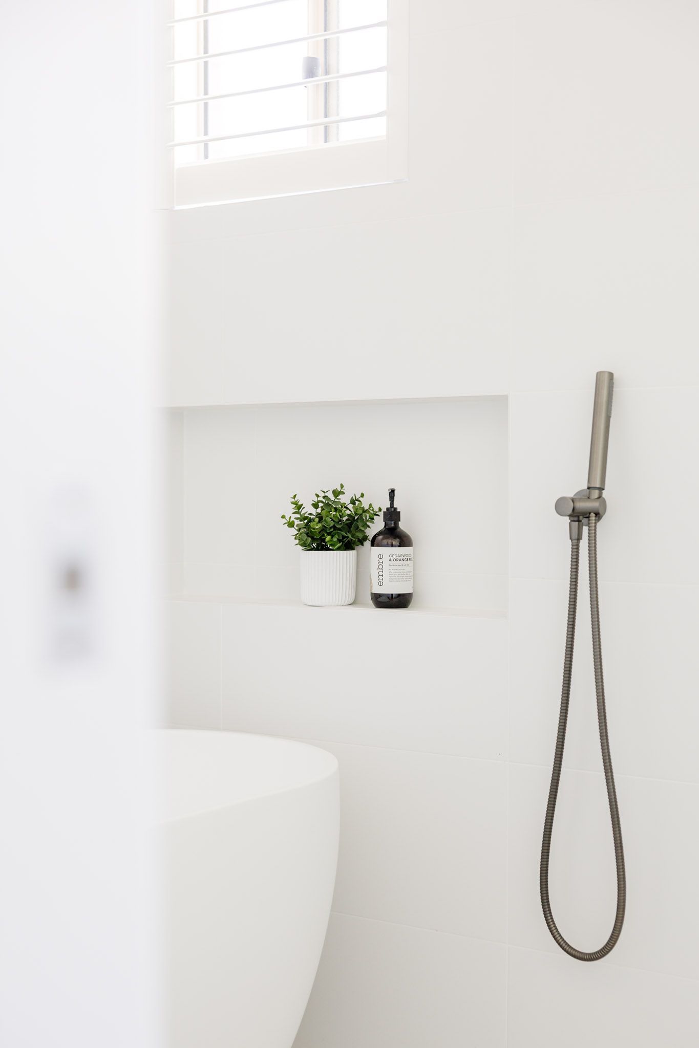 White bathroom with showerhead, built-in shelf with plant and bottle, and part of a tub.