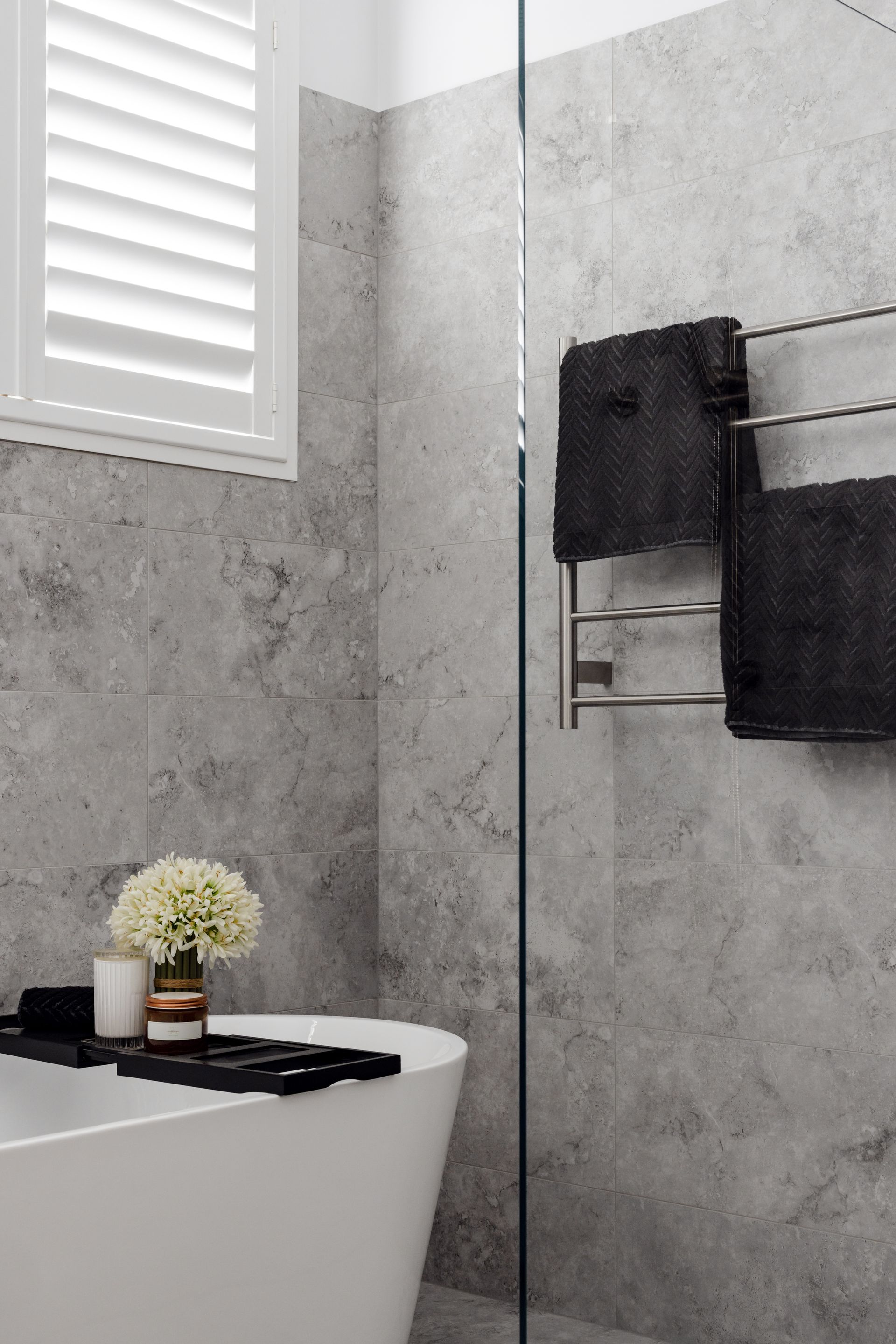 Bathroom with a white tub, gray tile, and black towels on a heated rack.