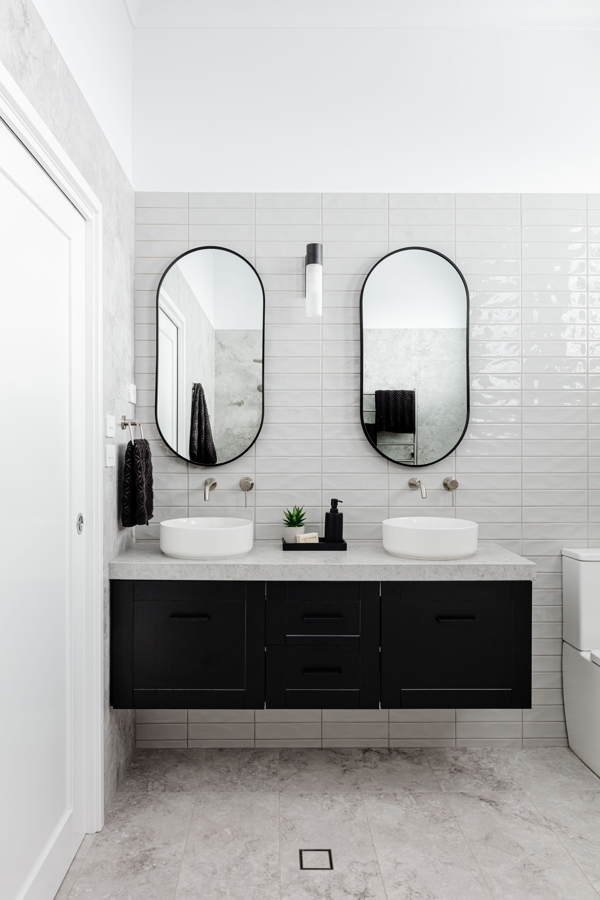 Modern bathroom with black vanity, white sinks, oval mirrors, and textured white tile.