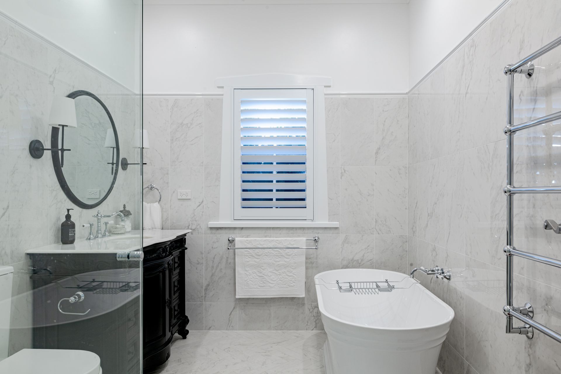 Elegant white bathroom with freestanding tub, dark vanity, and shuttered window.