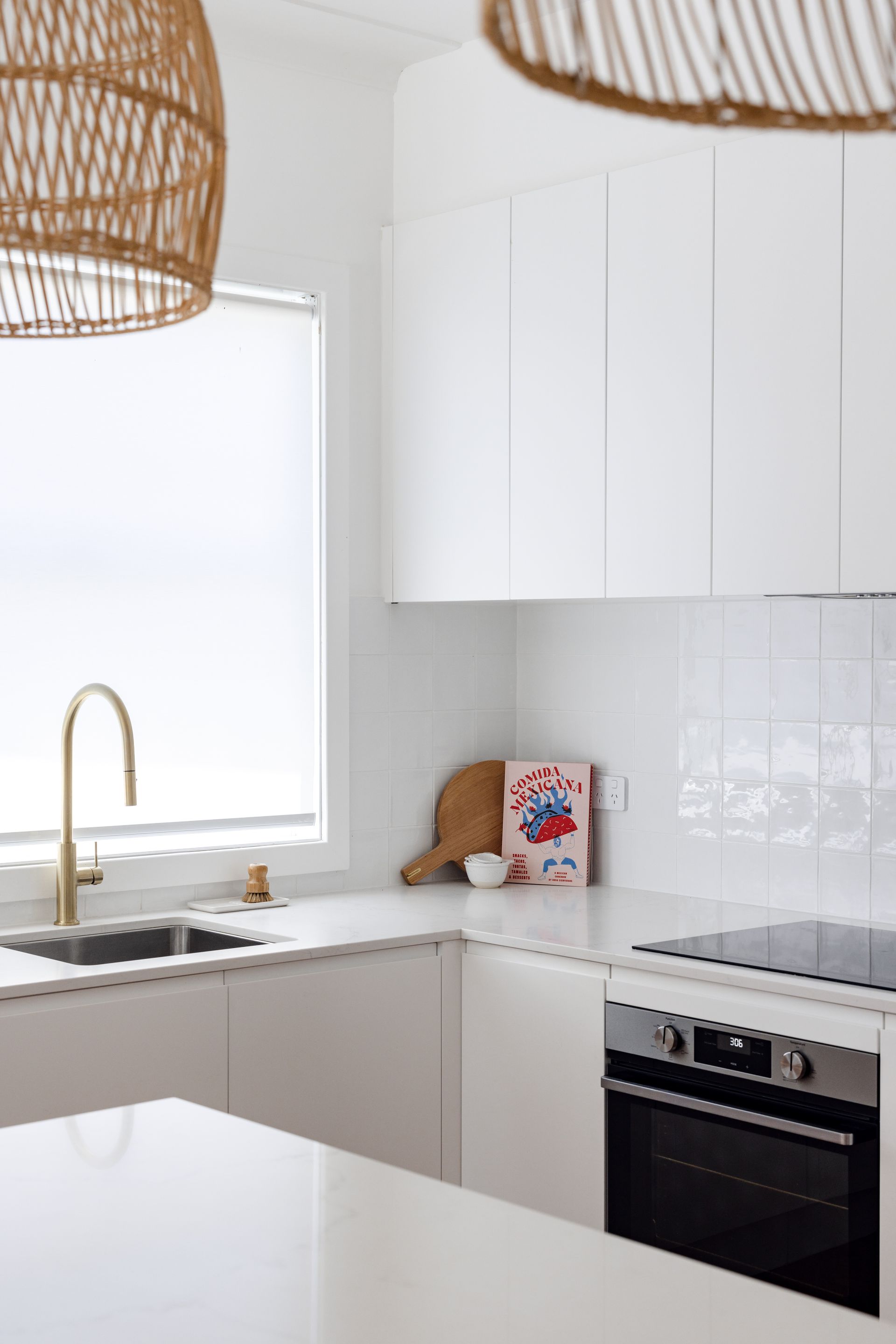 Modern white kitchen with gold faucet, wooden cutting board, and woven light fixtures.