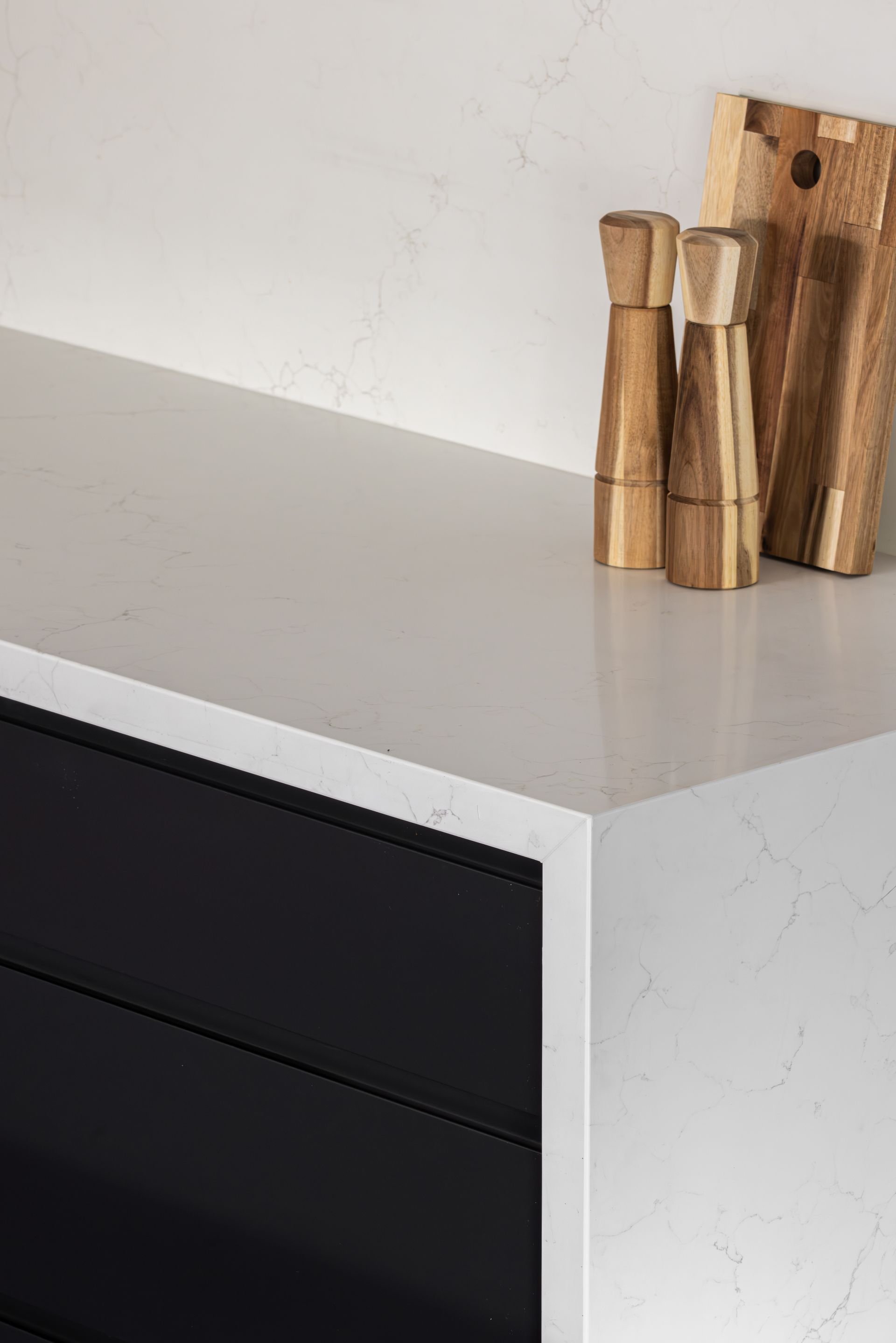 Close-up of a modern kitchen countertop: white surface, black drawers, wooden salt and pepper grinders, and a cutting board.