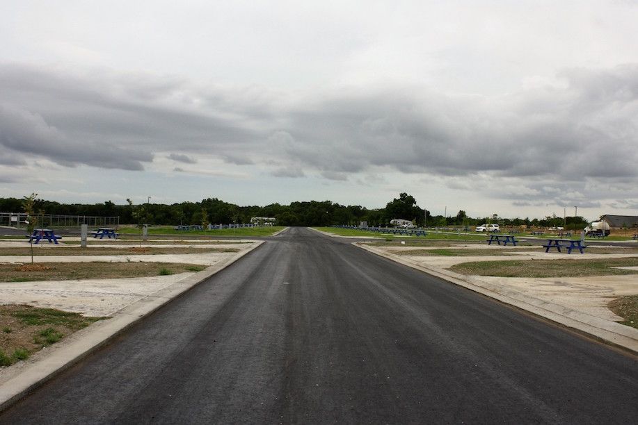 Paved road through an empty RV park under a cloudy sky. Picnic tables and trees line the road.