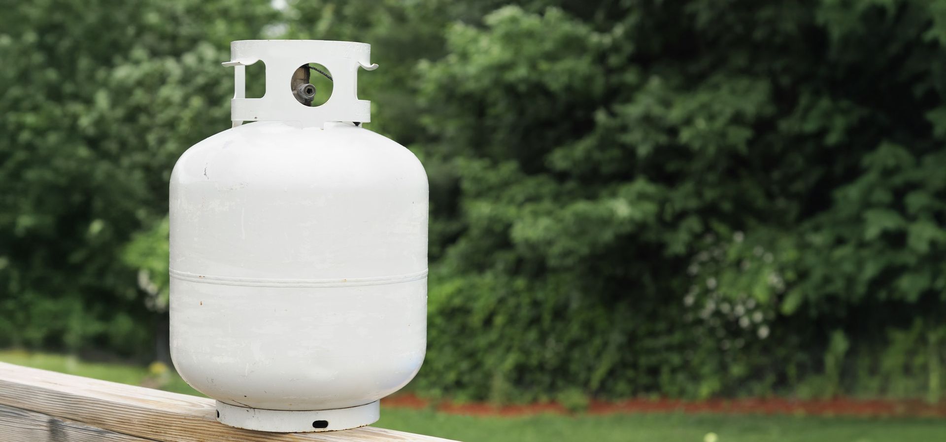 A white propane tank sitting outside, with green trees in the background.