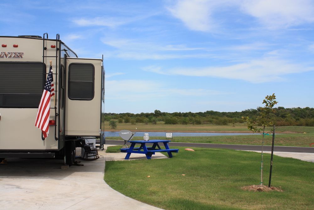 RV parked at campsite with American flag, picnic table, and view of water under blue sky.