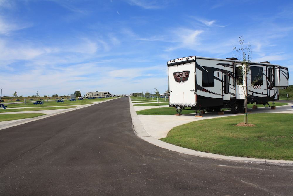 RV parked on grass beside paved road in a campsite. Blue sky, other RVs in distance.