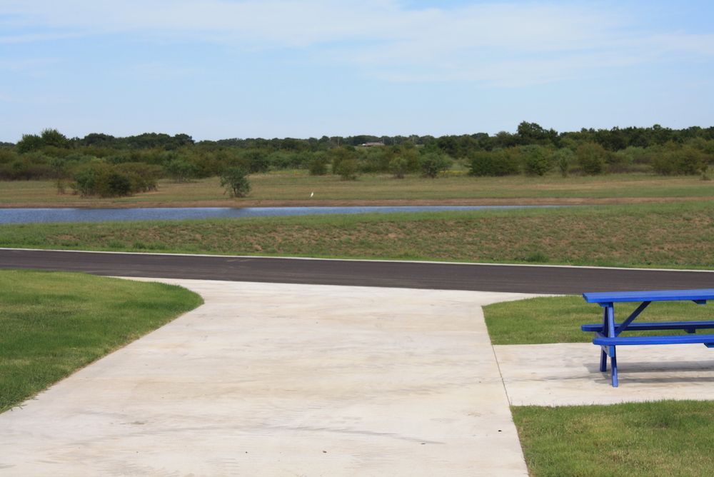 A picnic table on concrete near a grassy area, asphalt path, and a body of water.