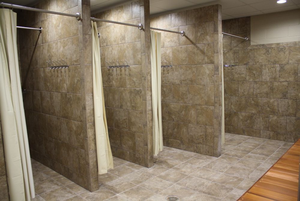 Public shower stalls with beige tiled walls, curtains, and a wooden bench.