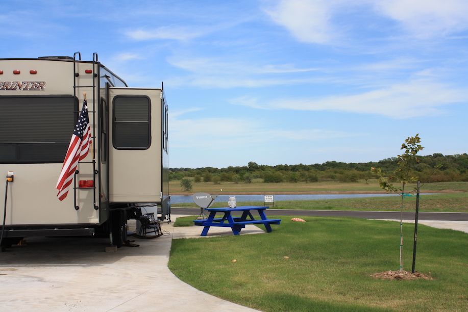 RV parked on concrete pad with a picnic table and American flag. Green grass, blue sky, and trees.