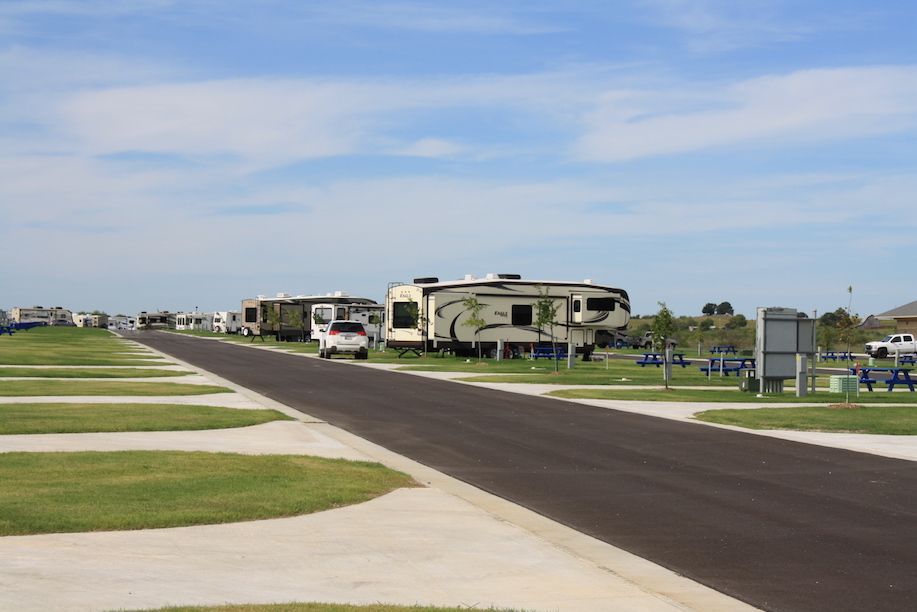 RV campground with a paved road, RVs parked on grass, blue picnic tables, and a cloudy sky.