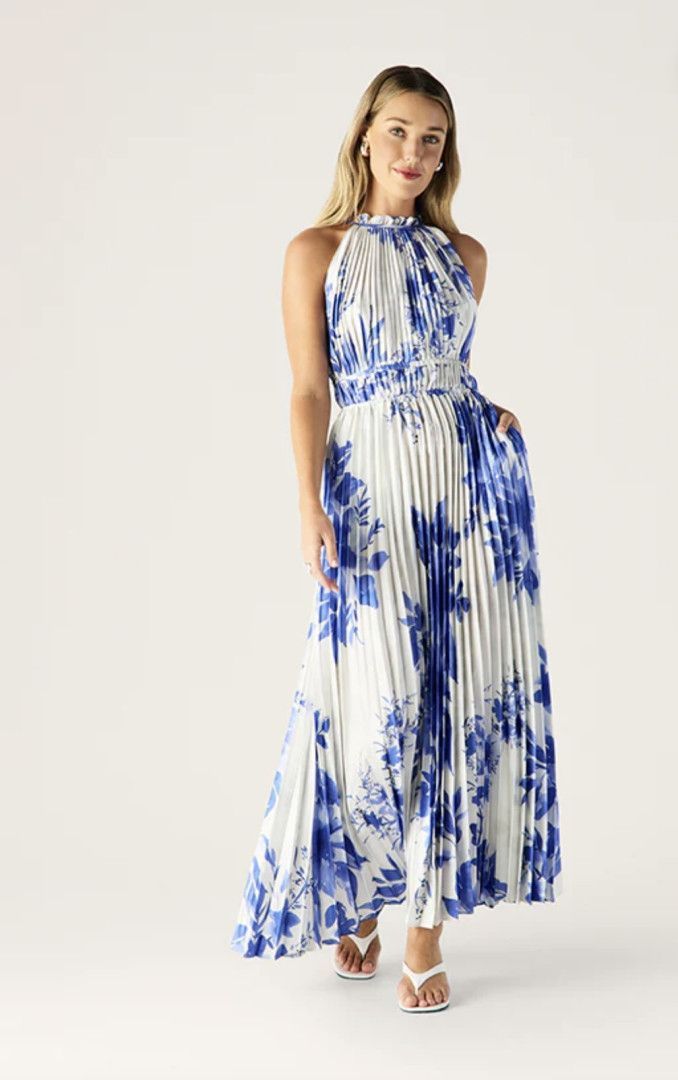 Woman in a white and blue floral pleated maxi dress, standing in a studio, smiling.