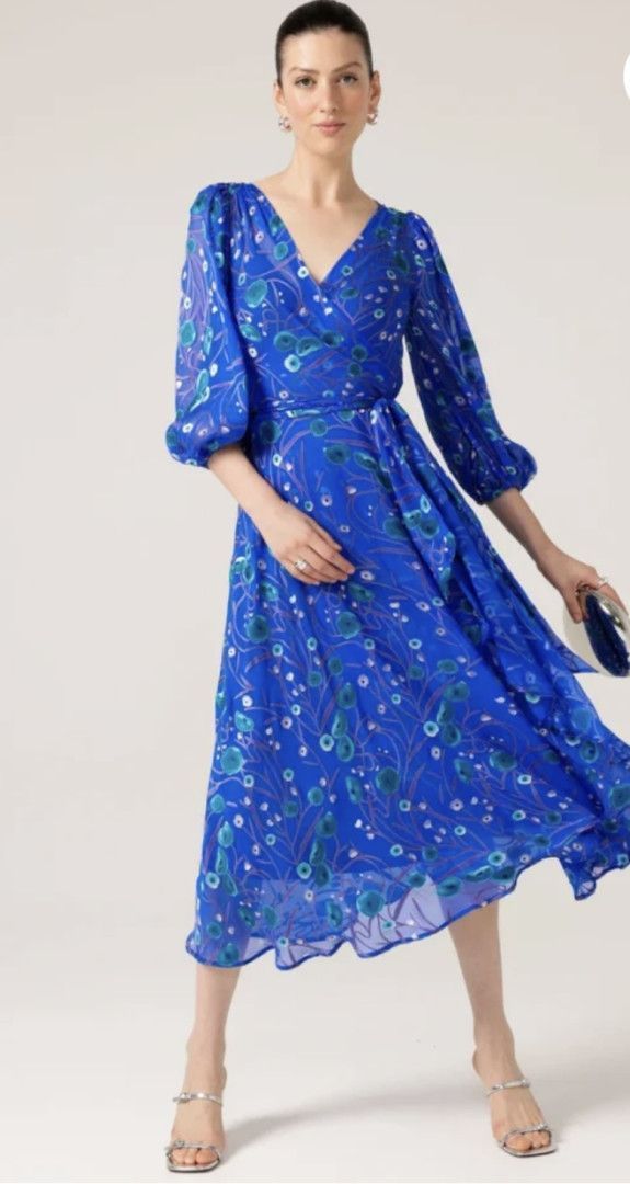 Woman in blue floral dress, looking at the viewer, with matching clutch and heels.