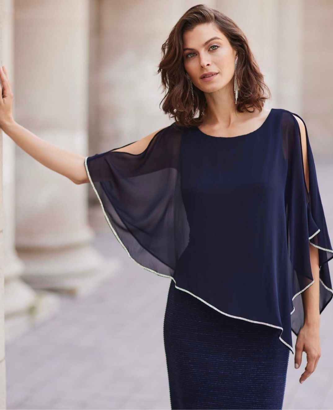 Woman in navy dress with sheer overlay, standing by a pillar, posing for the camera.