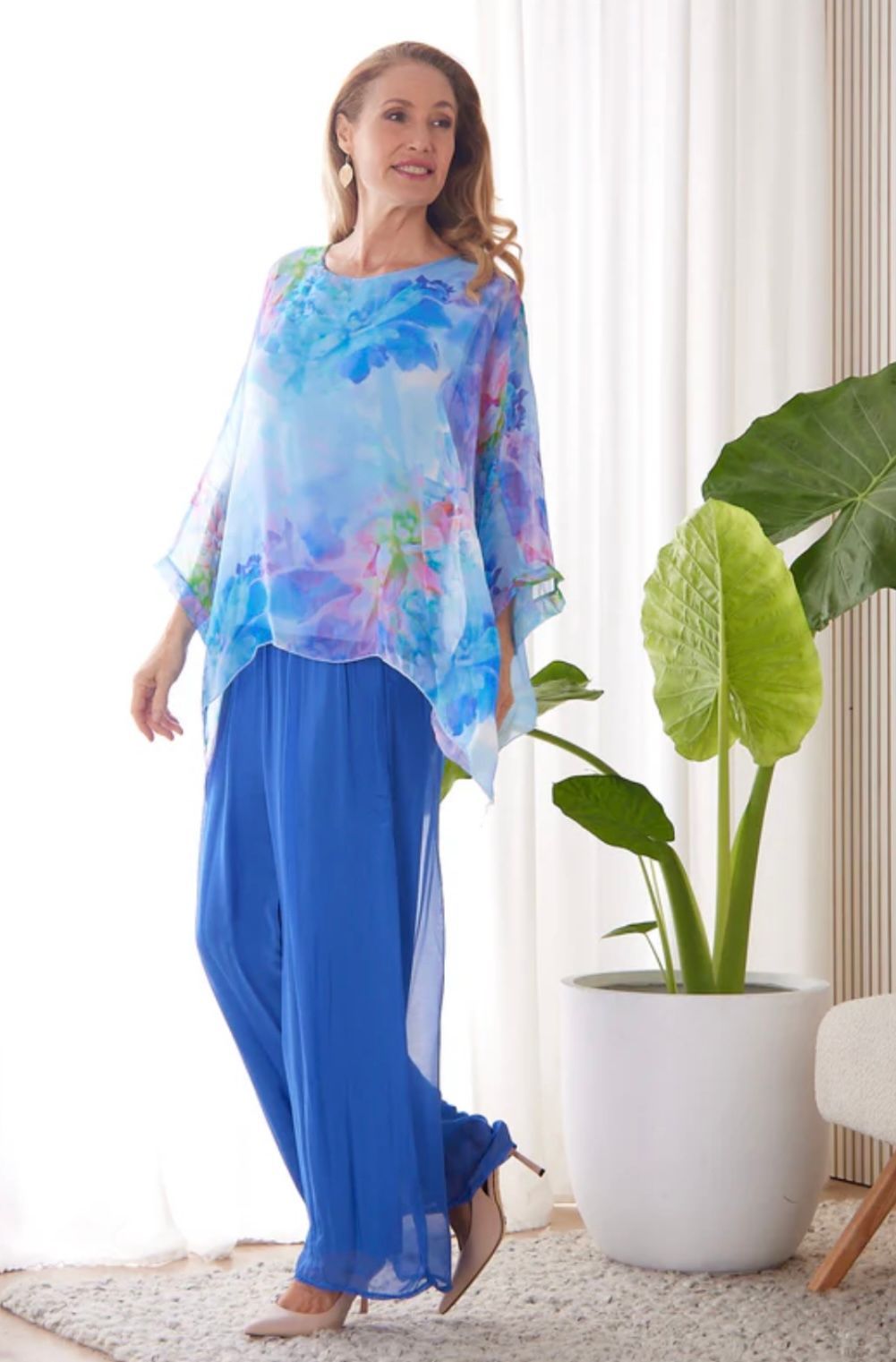 Woman wearing a blue floral tunic top and pants, standing indoors near a plant.