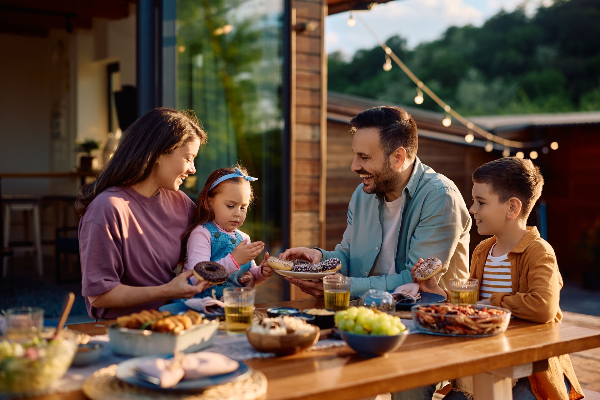 Family eating outdoors at a wooden table: woman, man, and two children sharing food.