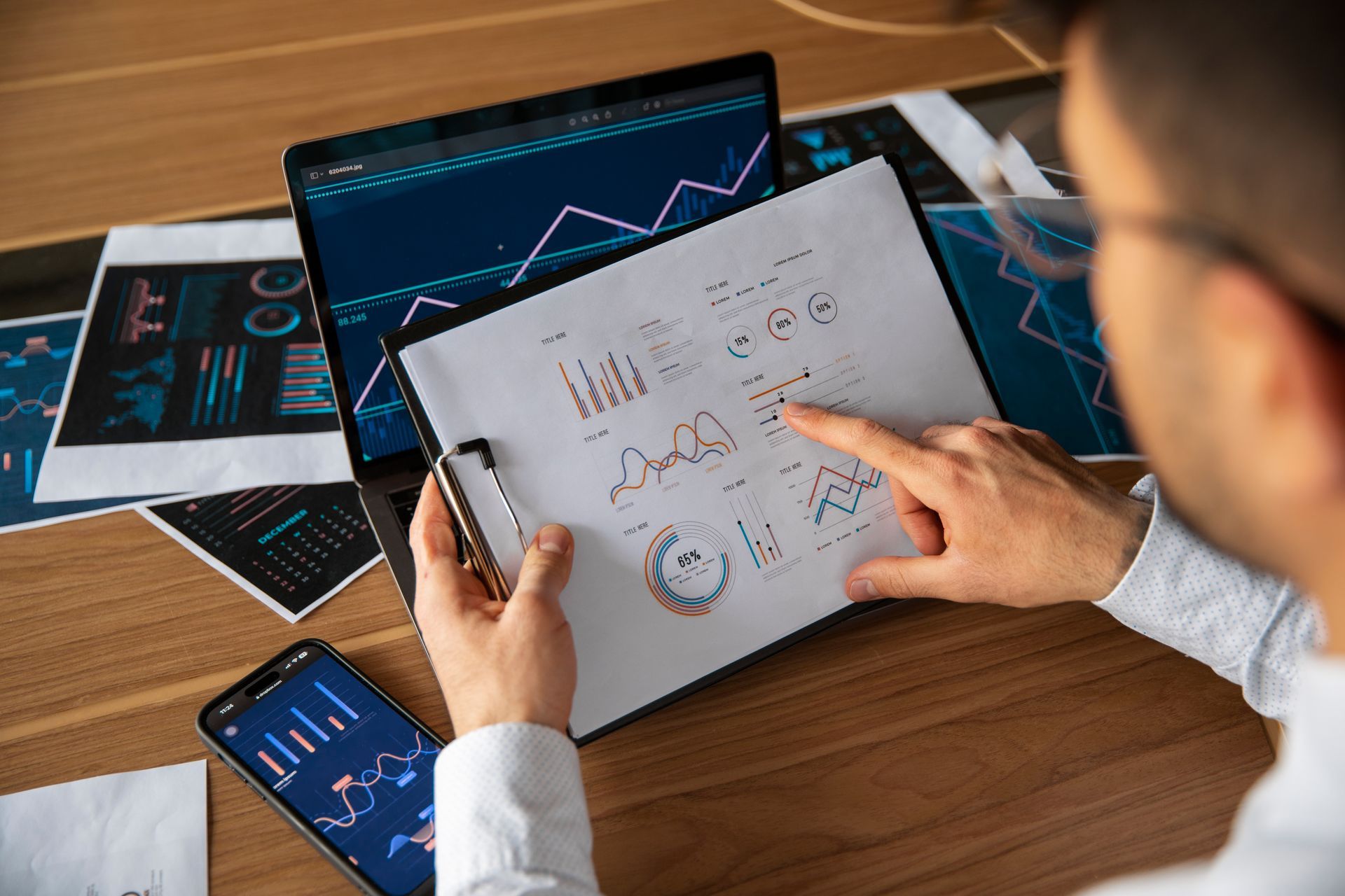 Person pointing at graphs on a clipboard, with a laptop and smartphone displaying charts on a wooden desk.