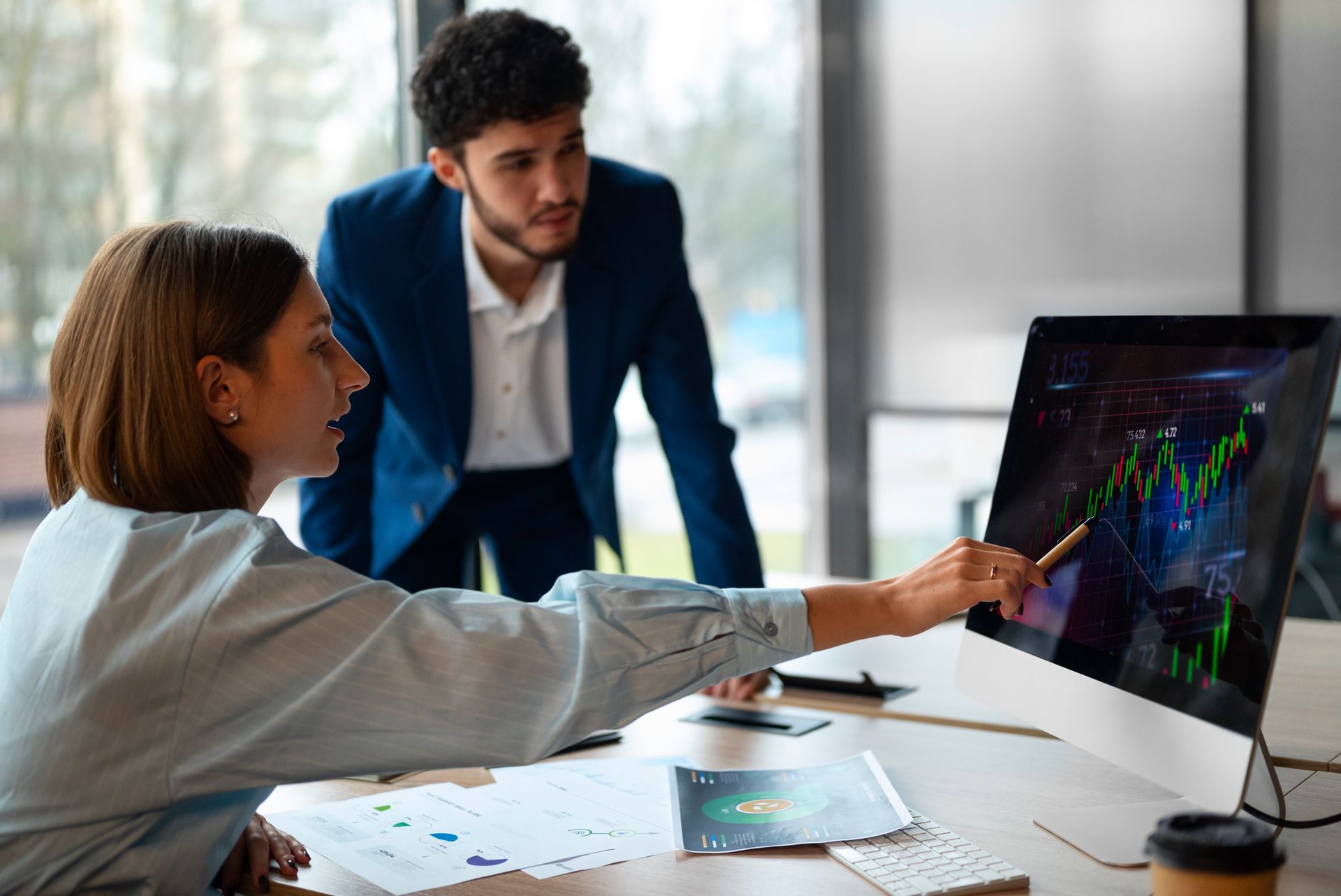 Woman points at graph on computer screen; man in suit looks on. In office.