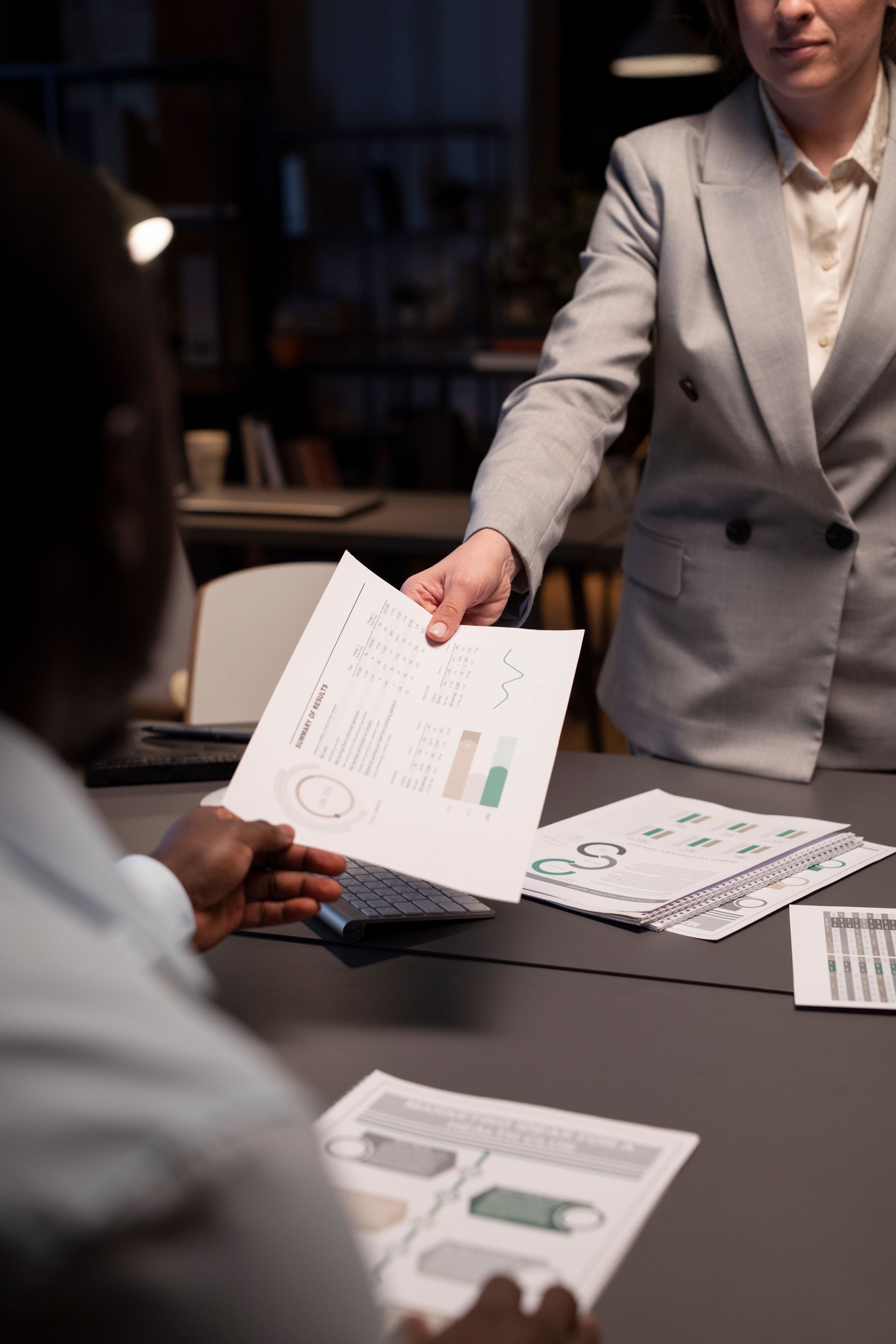 Person in a gray suit handing papers to another person at a table, documents scattered on the table.