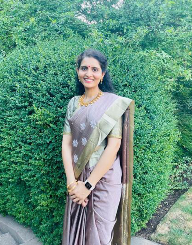 Woman in a lilac saree with gold accents, smiling, standing in front of a green hedge.