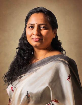 Woman in light-colored saree smiles at the camera, with long dark hair, against a brown background.