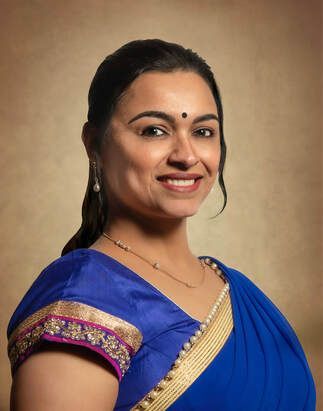 Woman with dark hair, wearing a blue sari with gold trim, smiling.