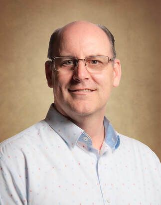 Man wearing glasses and a light blue patterned shirt smiling against a brown backdrop.