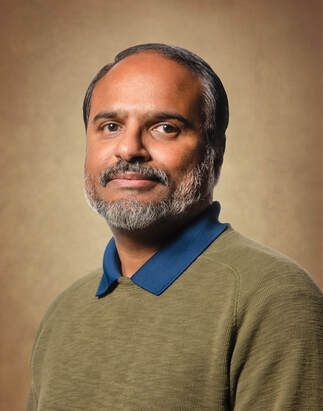 Man with dark hair and beard, wearing a green sweater with a blue collar, smiling.