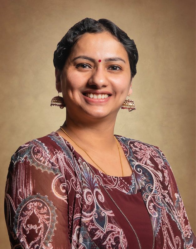 Woman with a bindi, wearing earrings and a printed top, smiling at the camera.