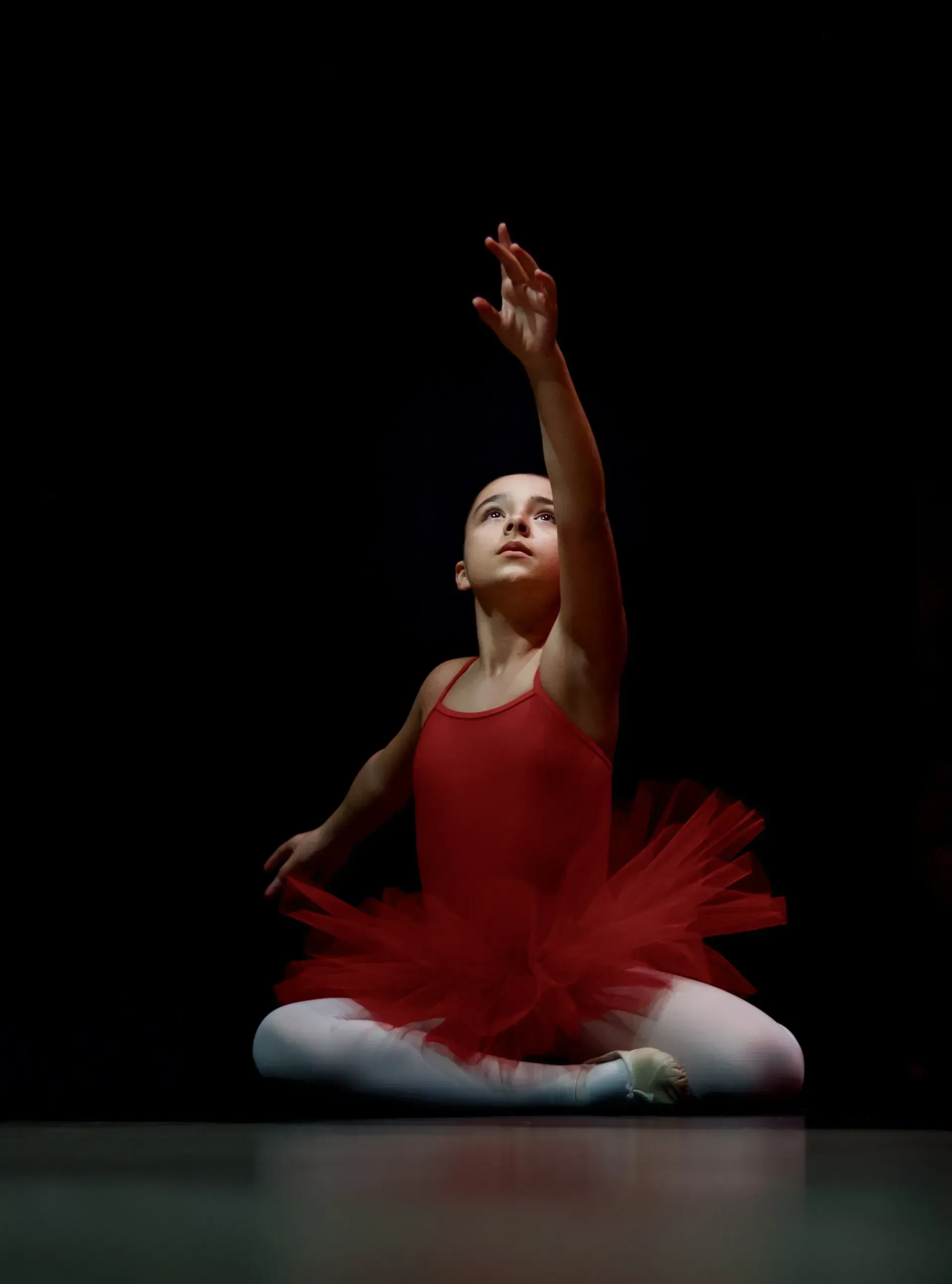 Young ballerina in red tutu, reaching upward, seated on floor, dark background.