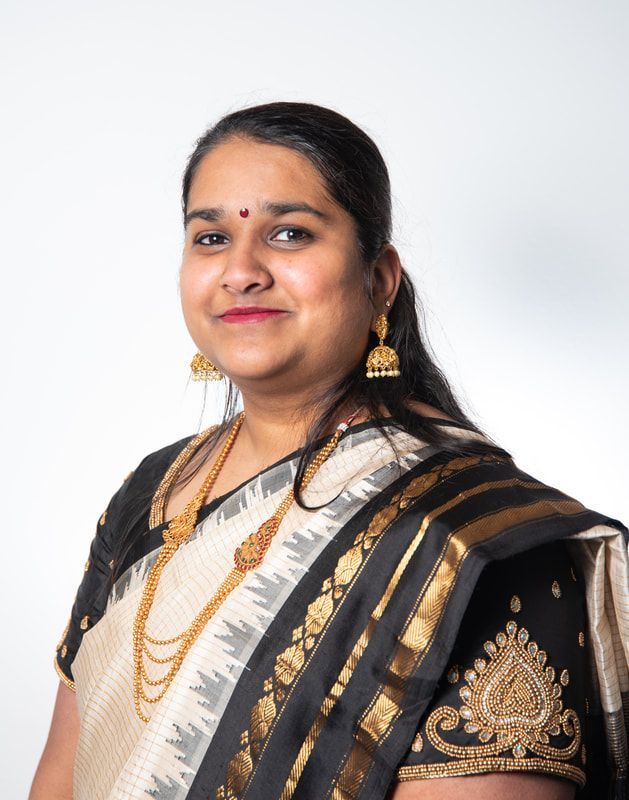 Woman in traditional attire, wearing gold jewelry, smiles, with a plain white background.